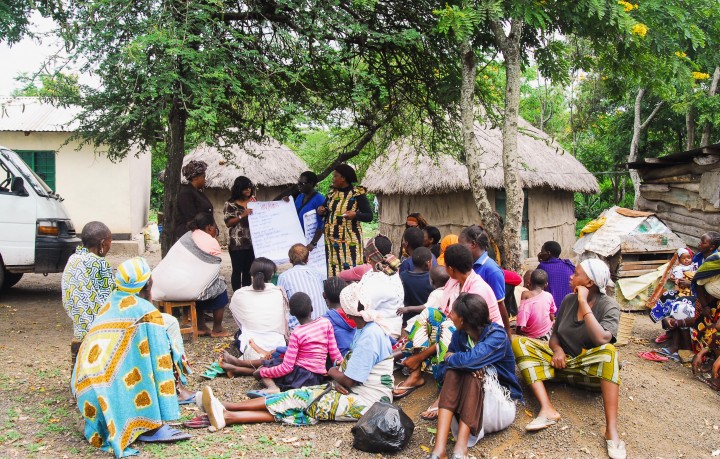 A group of people gather around a tree. They are watching the woman at the front who is writing in flip chart paper